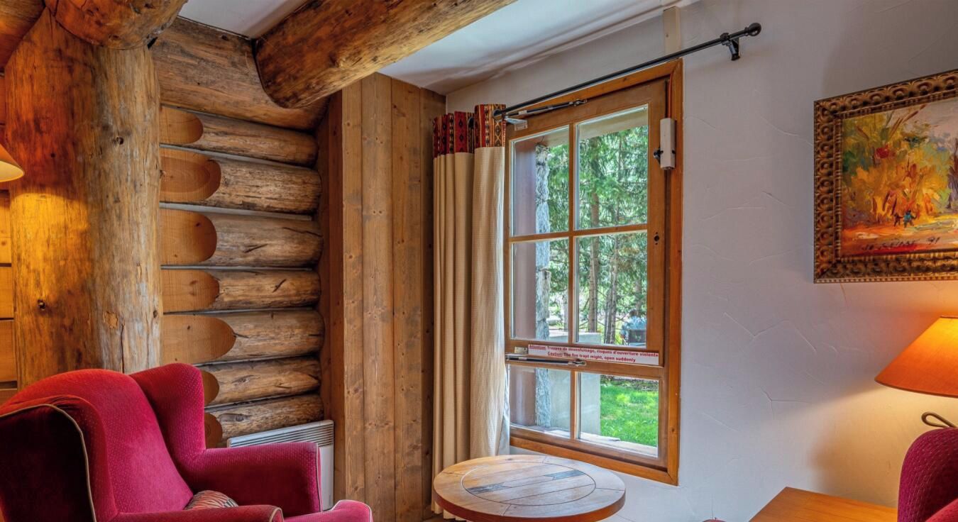 A log cabin interior with a red chair, a round wooden table, and a window with curtains.
