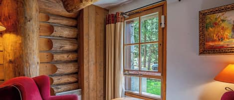 A log cabin interior with a red chair, a round wooden table, and a window with curtains.
