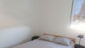 A single bed with a wooden frame and grey bedding, a small bedside table with a lamp, and a window with blinds. - Vrbo Property (Holtzheim)
