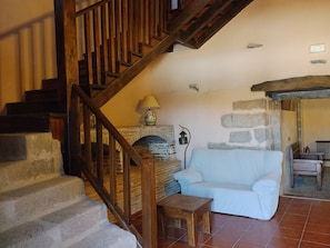 A stone fireplace, a wooden staircase, a white sofa, and a small wooden table in a room with stone walls and a tiled floor.