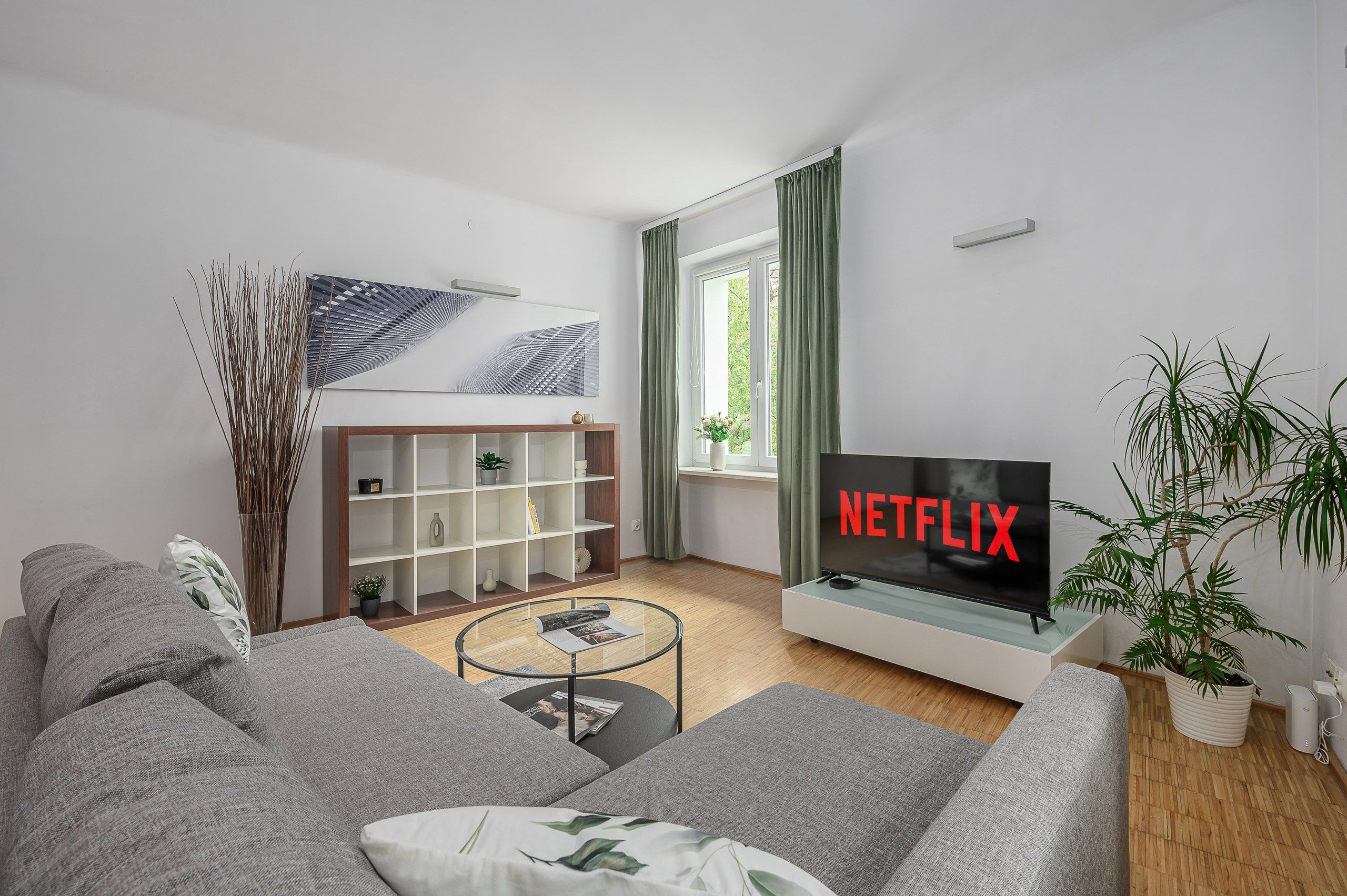 Un soggiorno moderno con un divano grigio, un tavolino da caffè, una libreria, un televisore che mostra 'NETFLIX' e una finestra con tende.