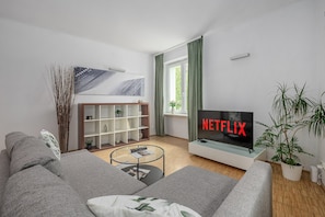 Una sala de estar moderna con un sofá gris, una mesa de centro, una estantería, un televisor mostrando 'NETFLIX' y una ventana con cortinas.