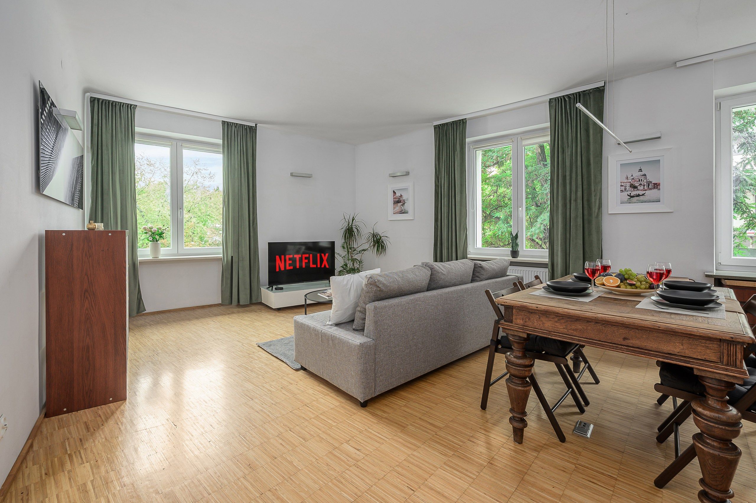 Un soggiorno moderno con un divano grigio, un tavolo da pranzo in legno e un televisore a schermo piatto che mostra 'NETFLIX'.