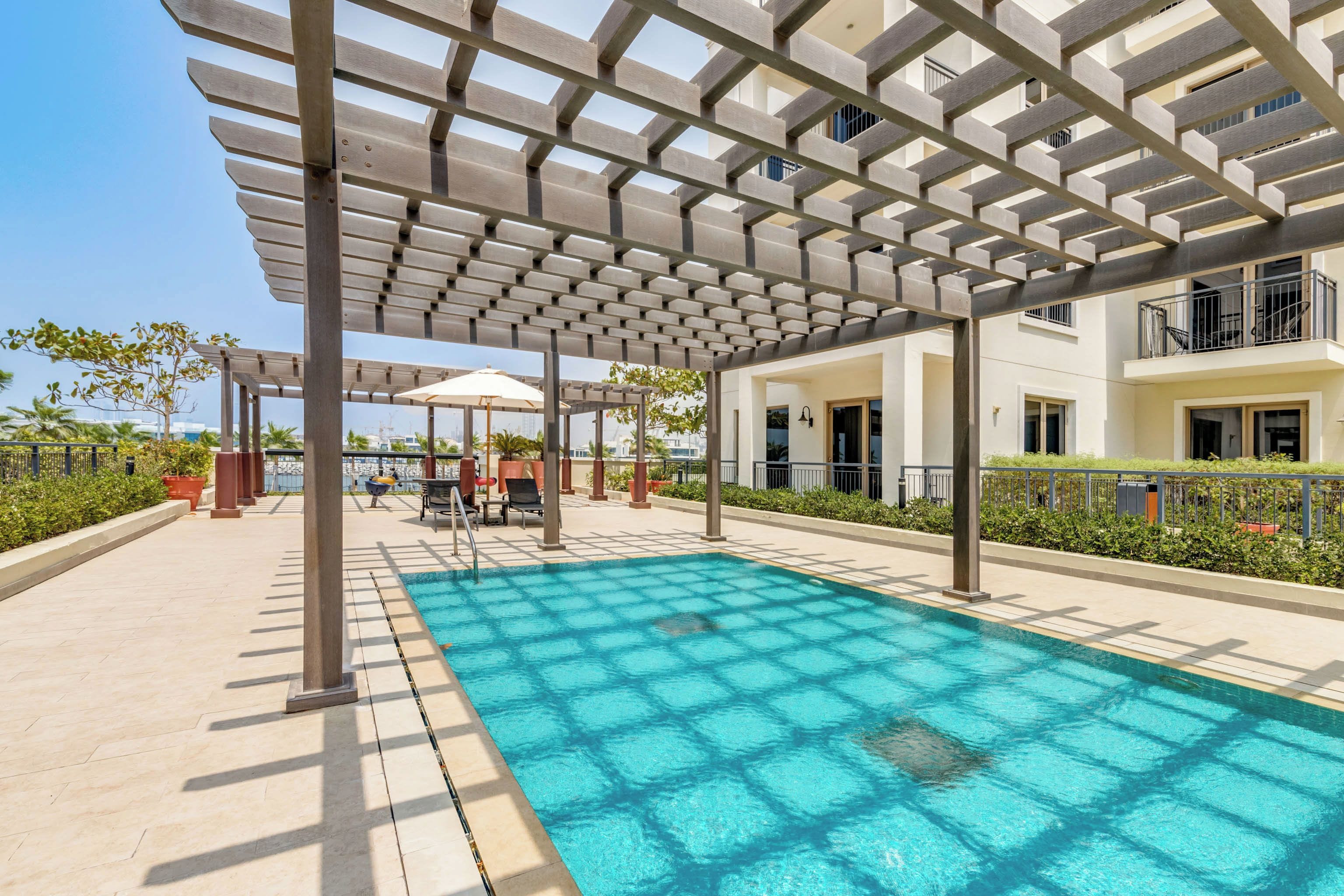 A swimming pool with a pergola structure overhead, surrounded by a landscaped area with a view of buildings in the background.