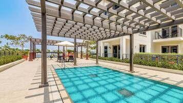 A swimming pool with a pergola structure overhead, surrounded by a landscaped area with a view of buildings in the background.