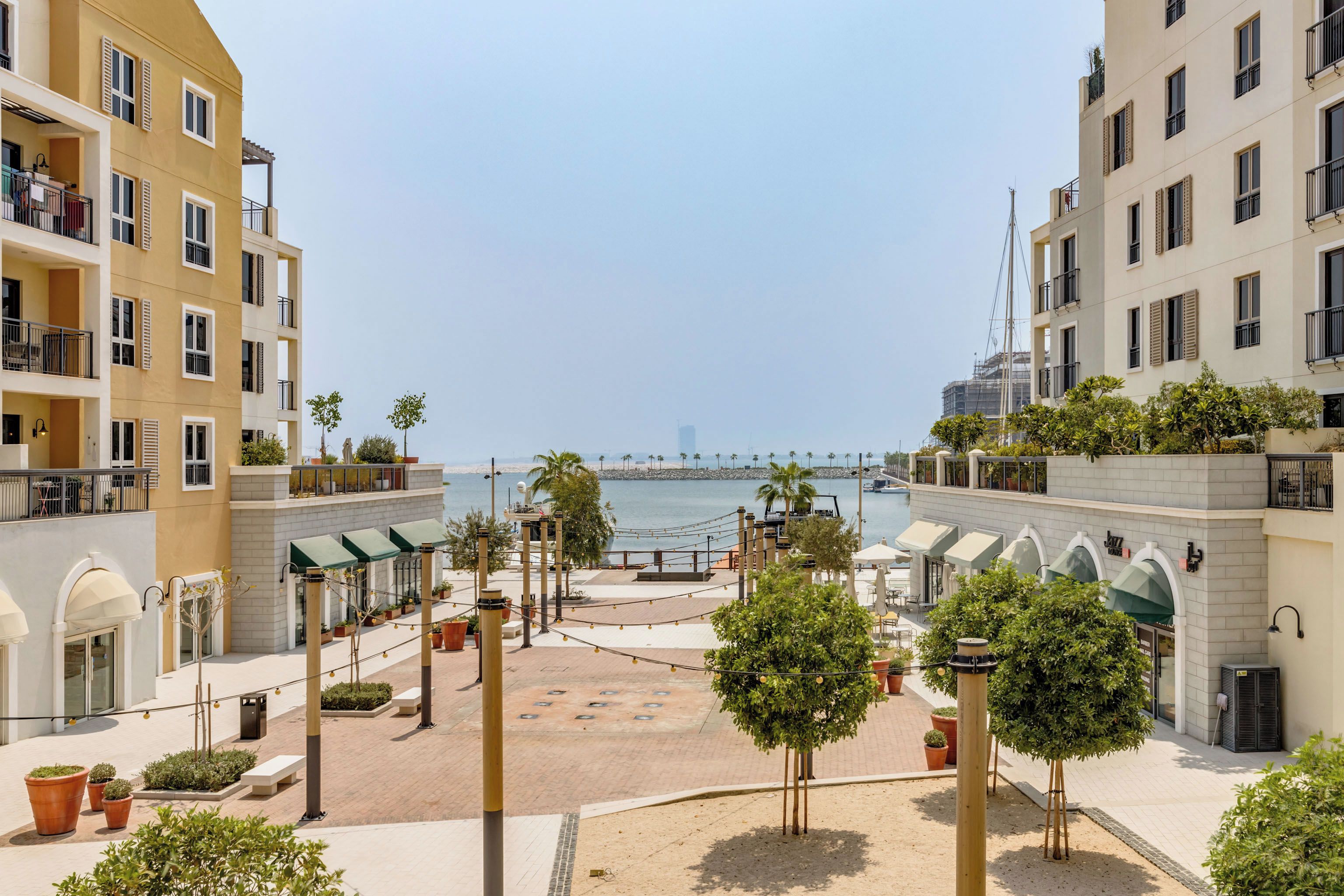 A coastal residential area with multiple-story buildings, a paved courtyard with seating, and a view of the sea.