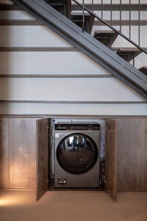 A compact, built-in washer and dryer unit in a modern apartment with a wooden finish and a staircase in the background.