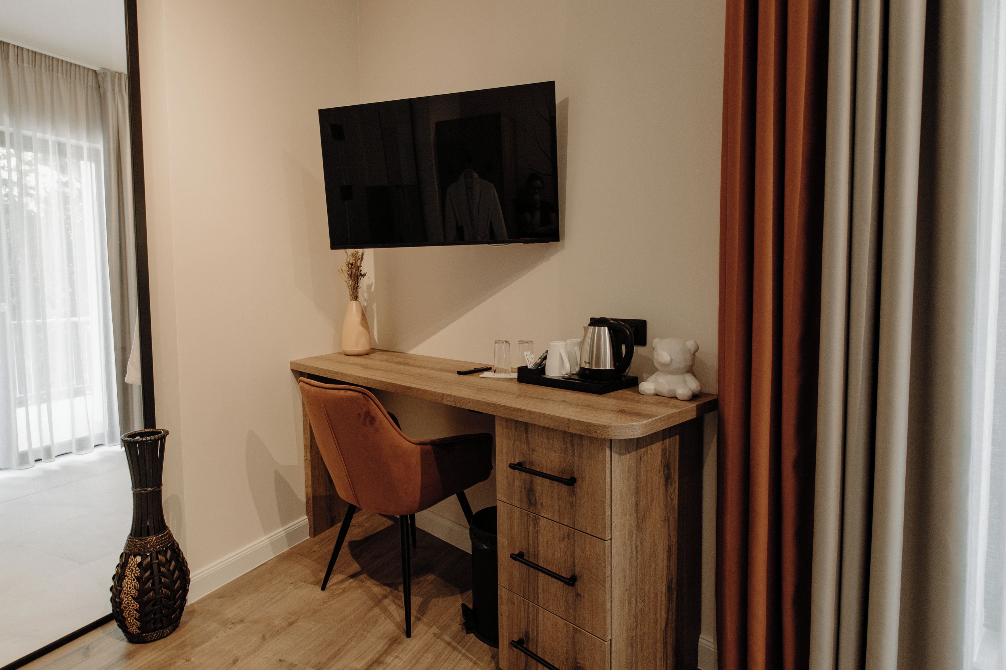 Una habitación pequeña y moderna con un escritorio de madera, una silla, un televisor fijado a la pared y una ventana con cortinas translúcidas.