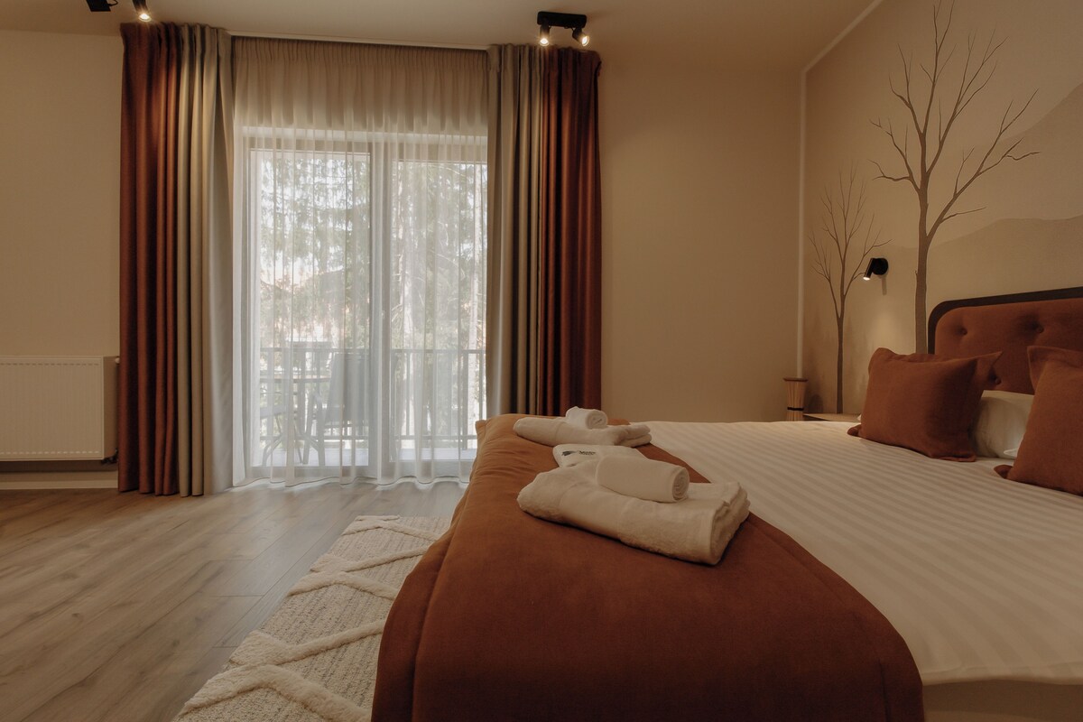 A bedroom with a large bed, a wooden floor, a window with curtains, and a wall-mounted lamp.