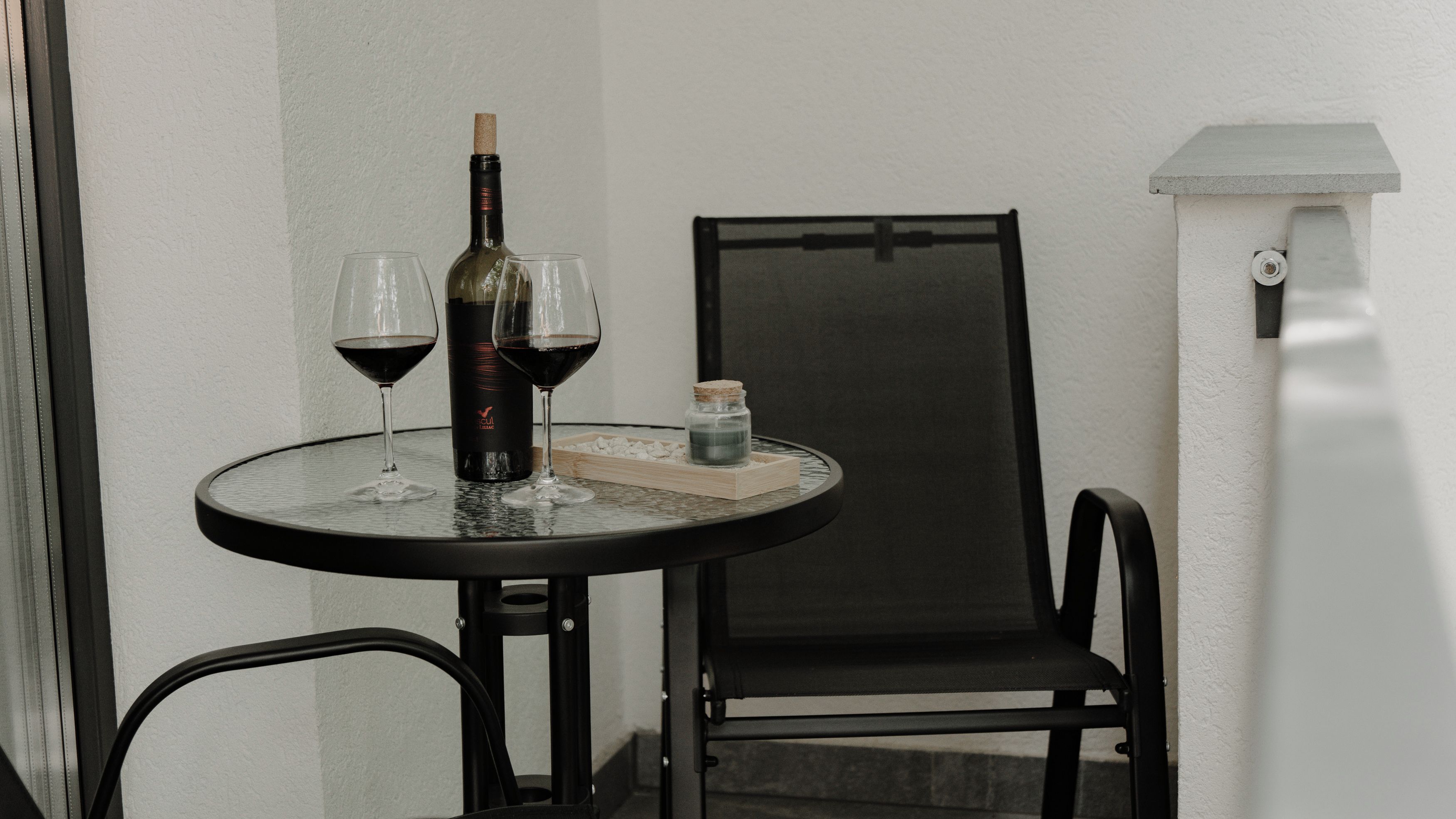 Una mesa redonda pequeña con una botella de vino, dos copas de vino tinto, una vela y un frasco pequeño sobre una superficie de vidrio.