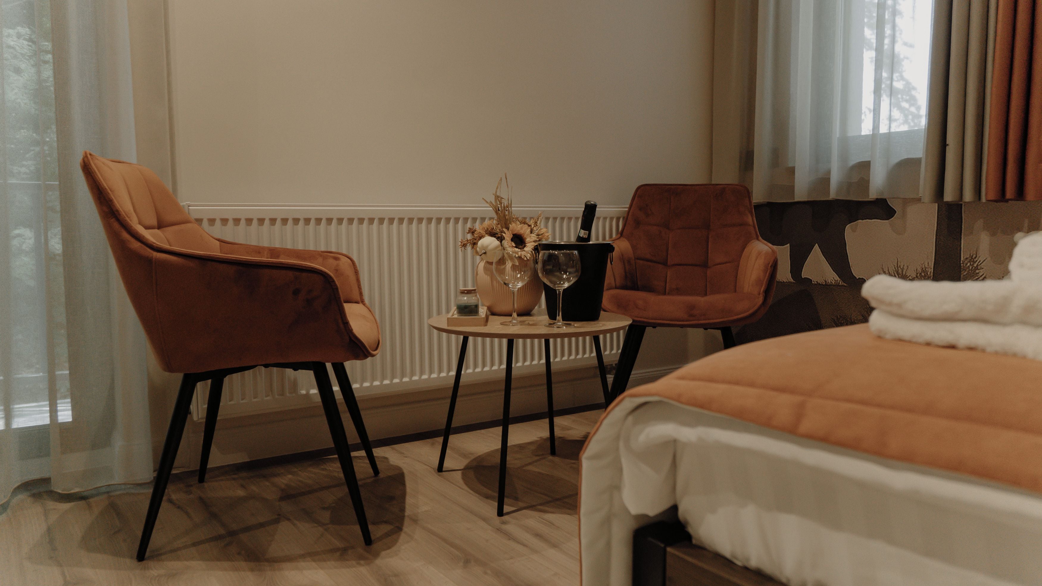 Une chambre d’hôtel comprenant un lit, deux fauteuils, une petite table avec un vase et des verres à vin, et un radiateur.
