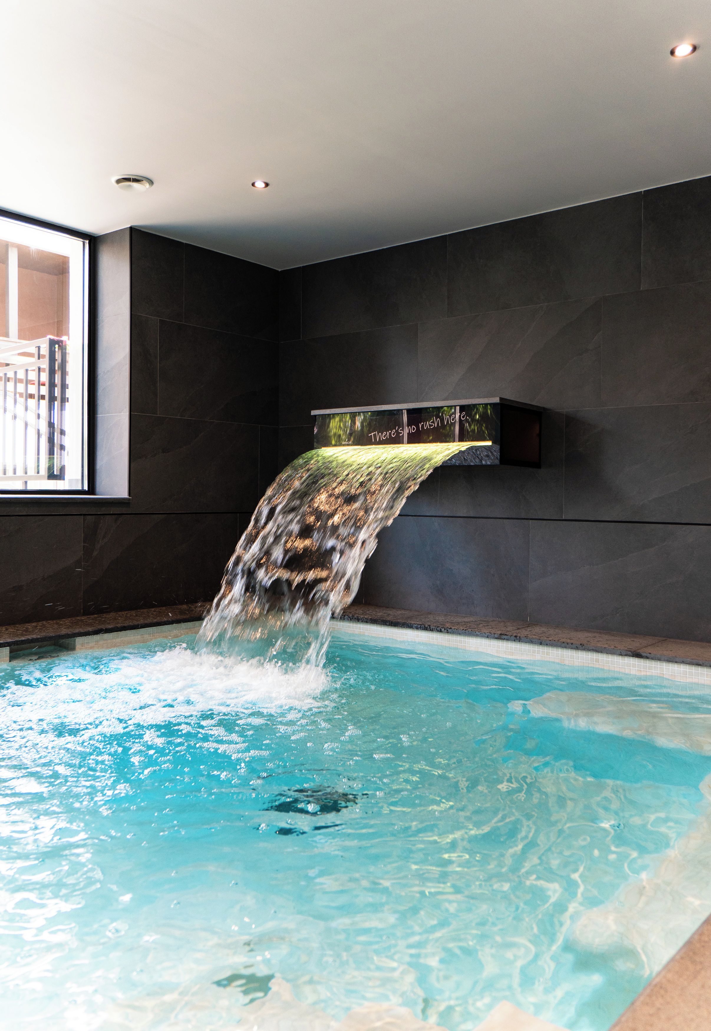 Une piscine agrémentée d’une cascade, avec des murs sombres et une fenêtre sur le côté gauche.