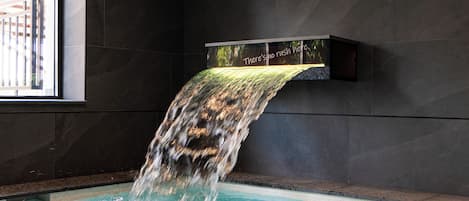 Piscina com cascata, paredes escuras e janela à esquerda.