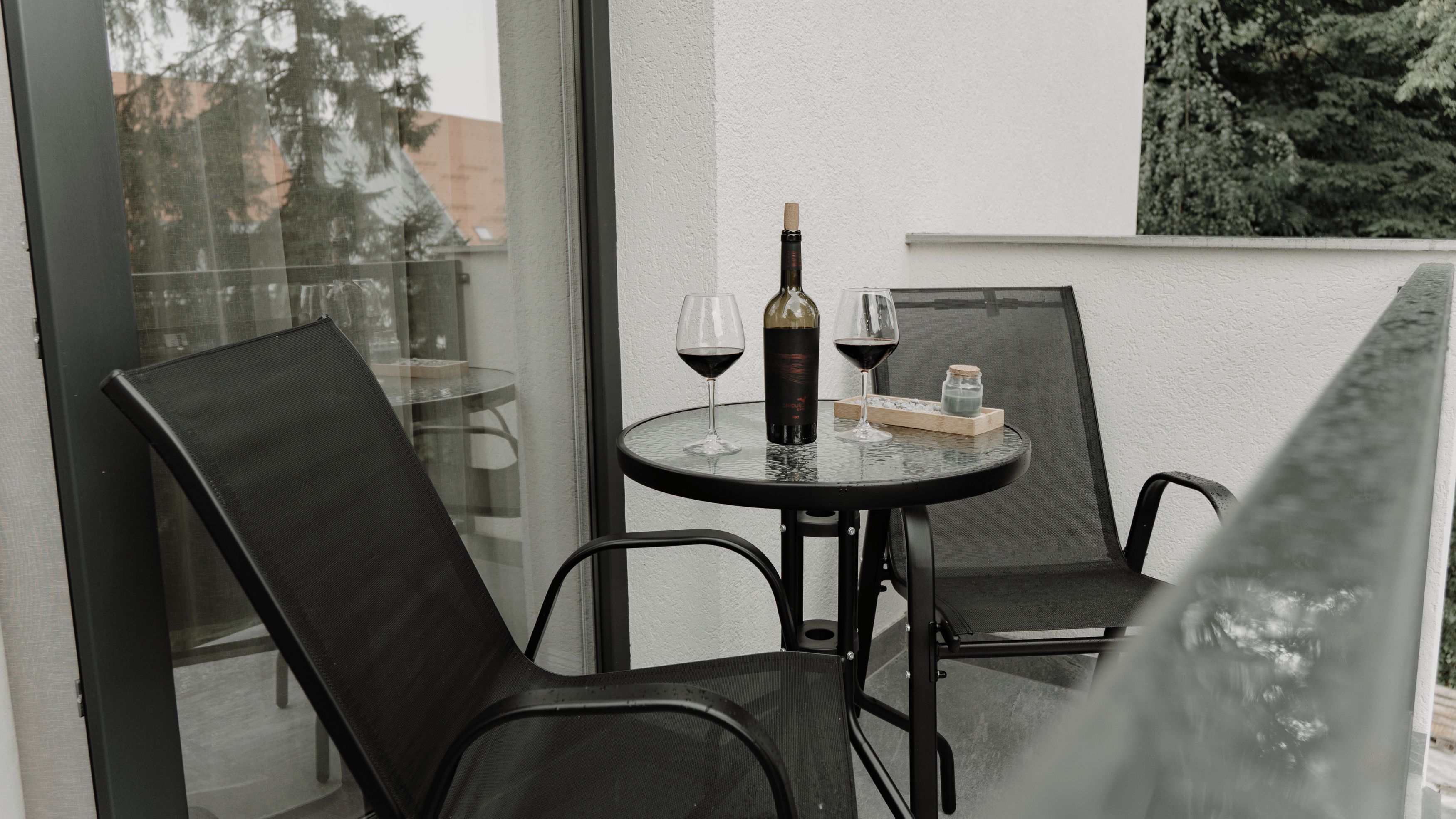 Una pequeña mesa redonda con una botella de vino y dos copas en un balcón con sillas negras.