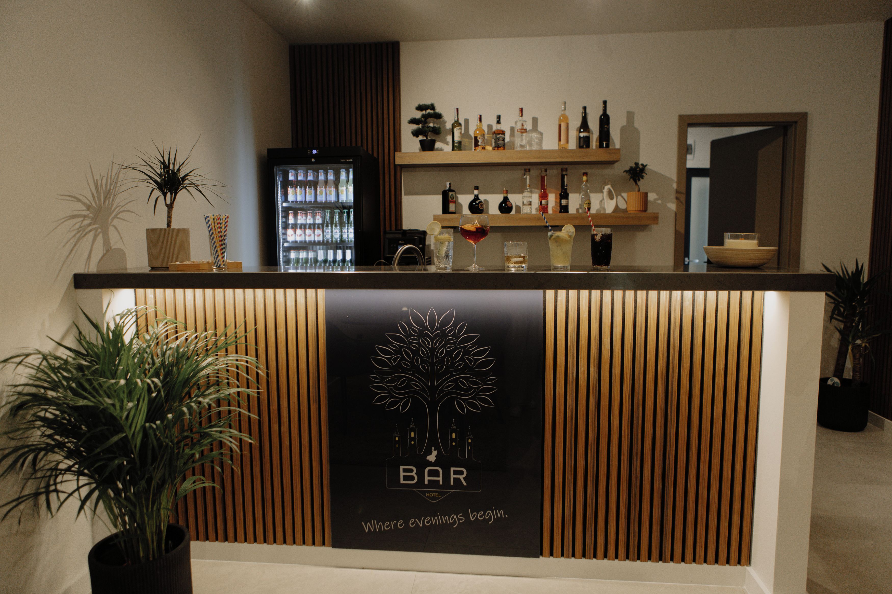 Zona de bar con mostrador de madera, un refrigerador lleno de bebidas y un cartel que dice 'BAR Donde comienzan las noches'.