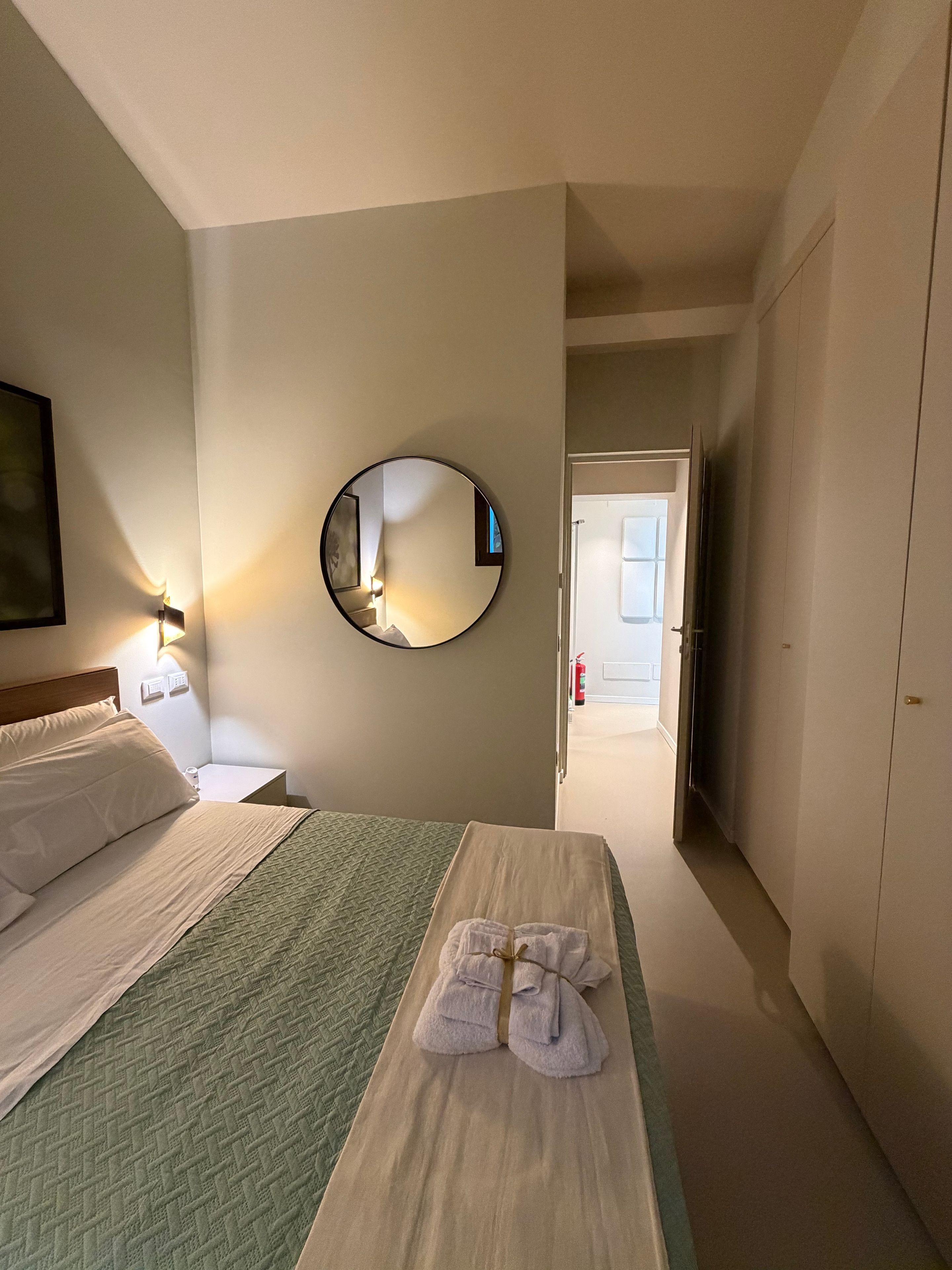 Hotellihuone, jossa on sänky, yöpöytä, peili, pyyheteline ja ovi, joka johtaa toiseen huoneeseen.