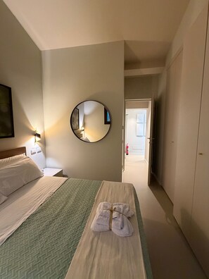 A hotel room with a bed, a bedside table, a mirror, a towel rack, and a door leading to another room.