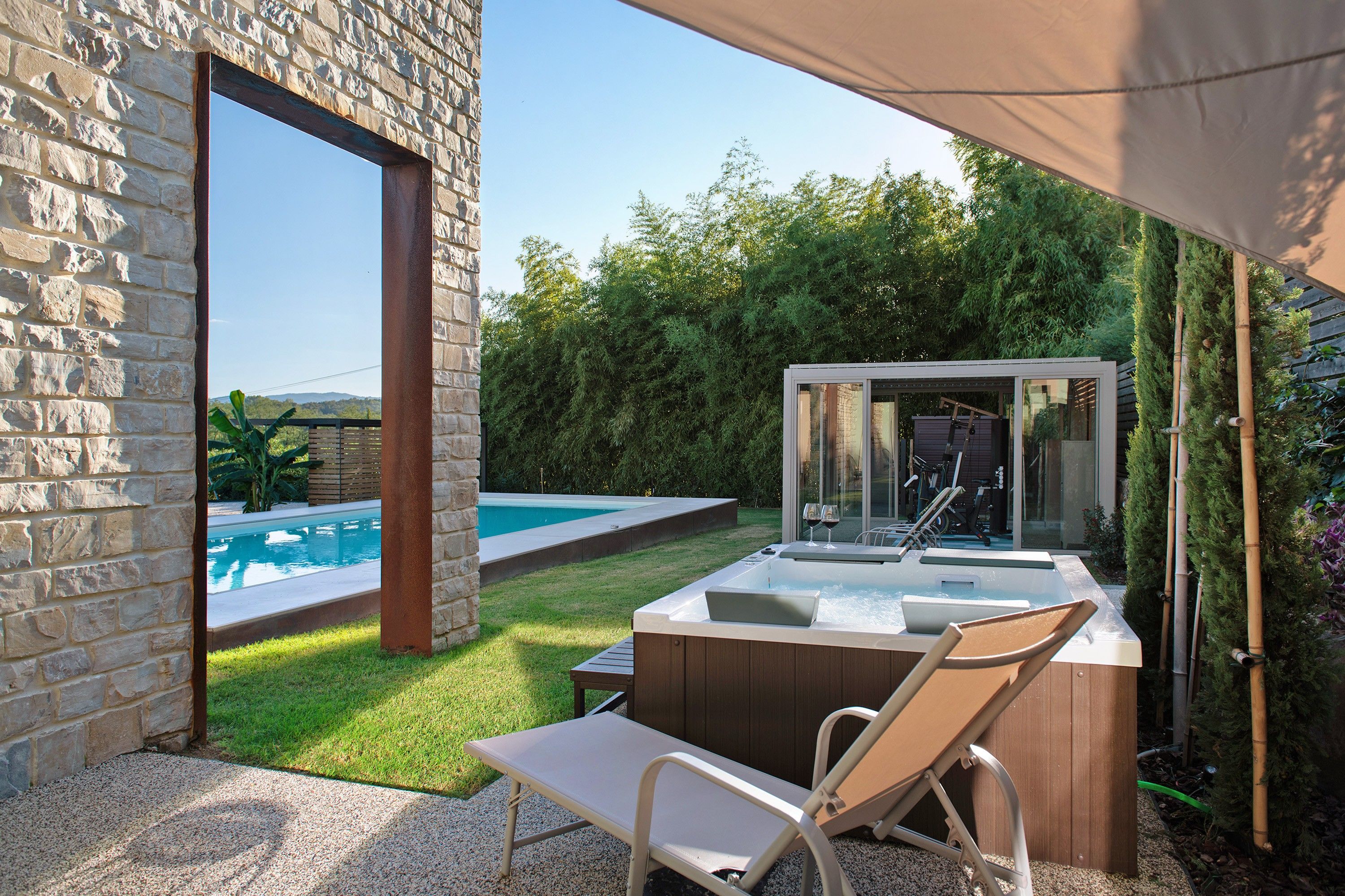 Un muro in pietra, una piscina, una vasca idromassaggio e una sedia a sdraio in un giardino.