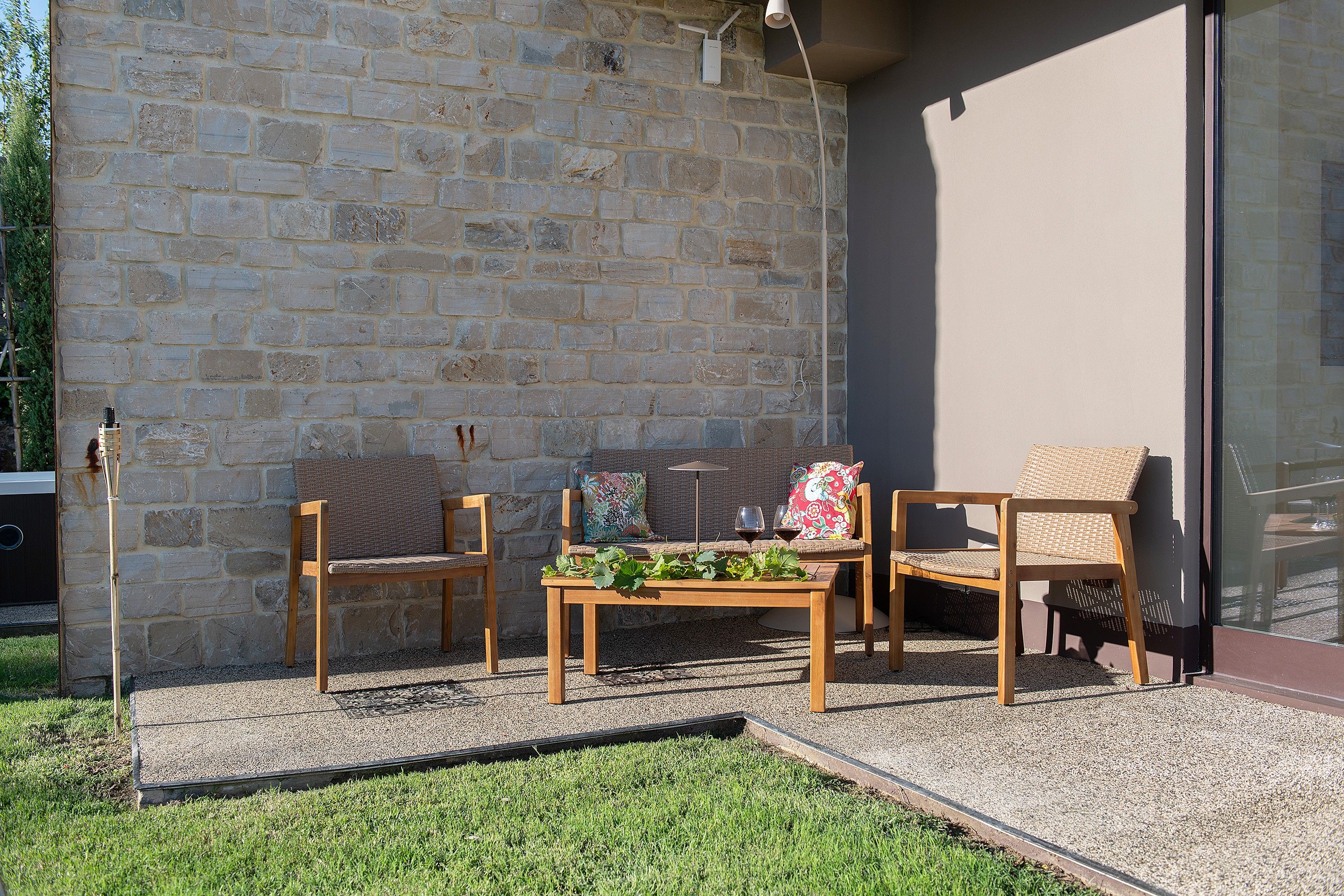 An outdoor seating area with two wooden chairs and a table, a stone wall, and a glass door.