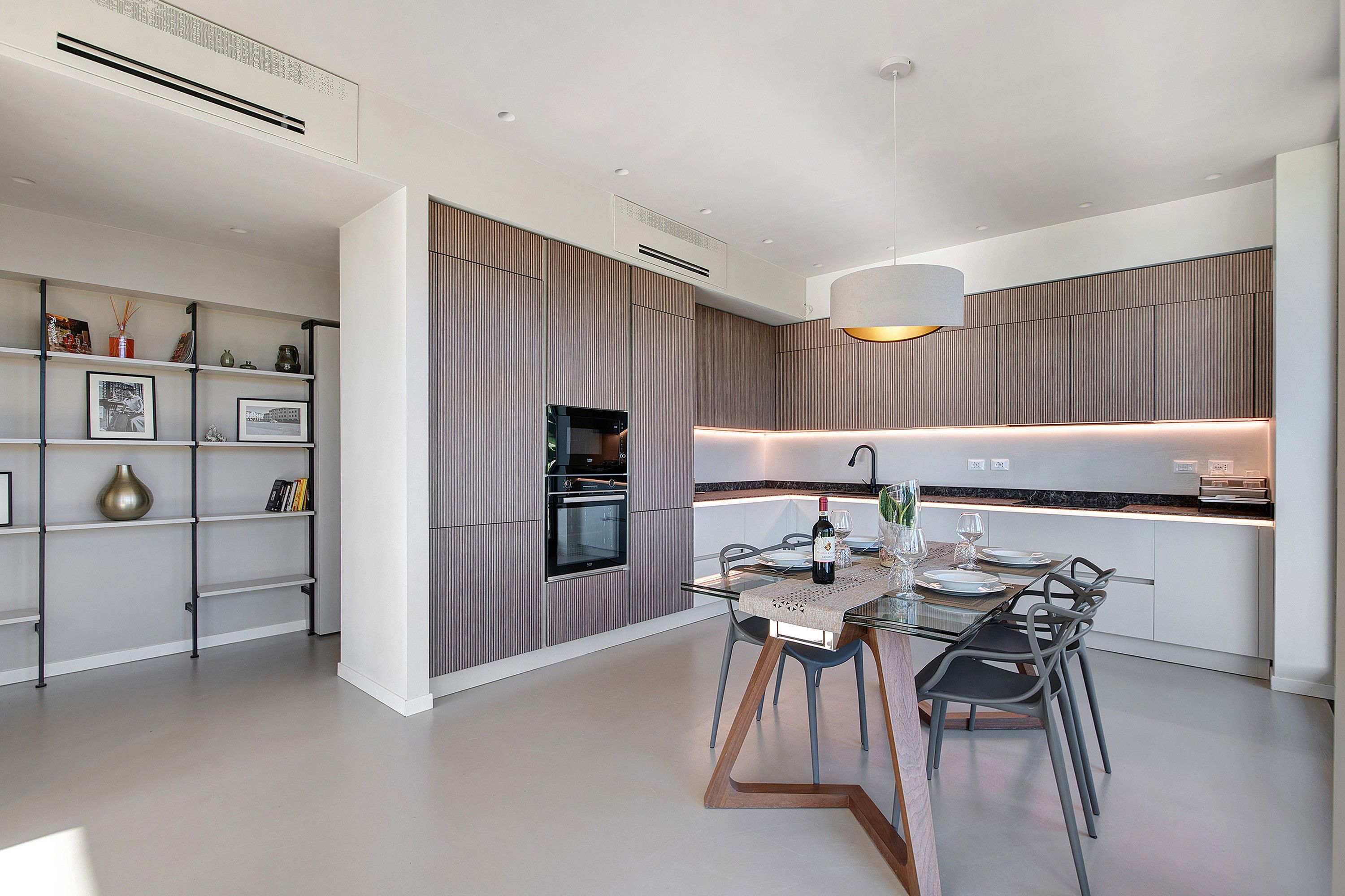 A modern kitchen with built-in appliances, a dining area with a glass table, and a decorative shelf with books and vases.
