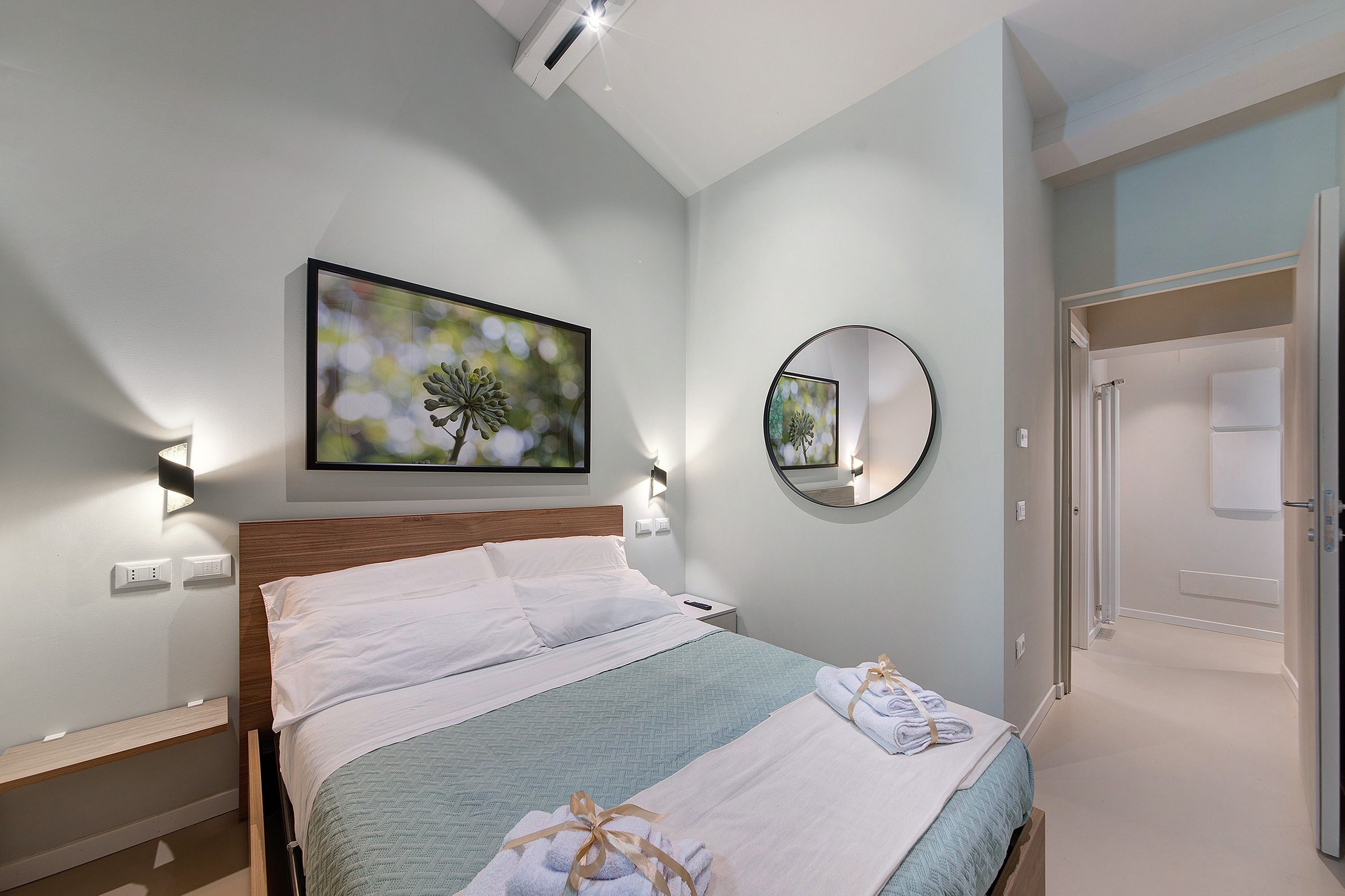 A hotel room with a bed, a wall-mounted TV, a round mirror, and a door leading to another room.