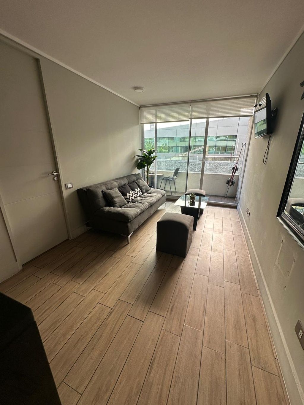 Een moderne woonkamer met een bank, een poef en een groot raam met uitzicht op een gebouw.
