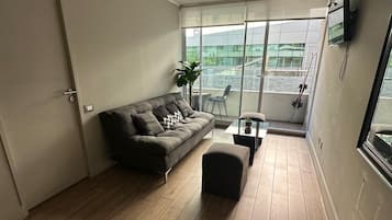 Een moderne woonkamer met een bank, een poef en een groot raam met uitzicht op een gebouw.