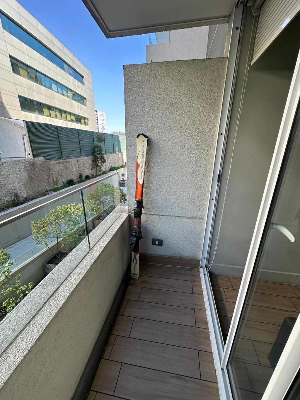 Un balcón con una tabla de snowboard apoyada en la pared, un vidrio y vista de un edificio al otro lado de la calle.