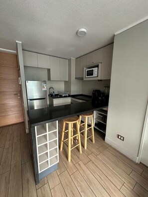 A compact kitchen with a black countertop, stainless steel appliances, wooden stools, and a microwave.