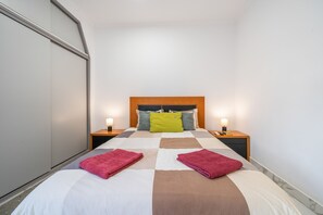 Un lit bien fait, recouvert d’une couverture à carreaux, avec deux oreillers et deux serviettes pliées. Il y a deux tables de chevet, chacune avec une lampe, et une télécommande est posée sur l’une d’elles.