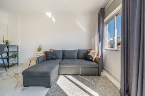 A modern living room with a grey sectional sofa, a small side table, a potted plant, and a view of buildings through a window with curtains.