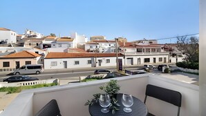 Un balcon avec une table, deux chaises et une vue sur une rue bordée d’immeubles et de voitures garées.