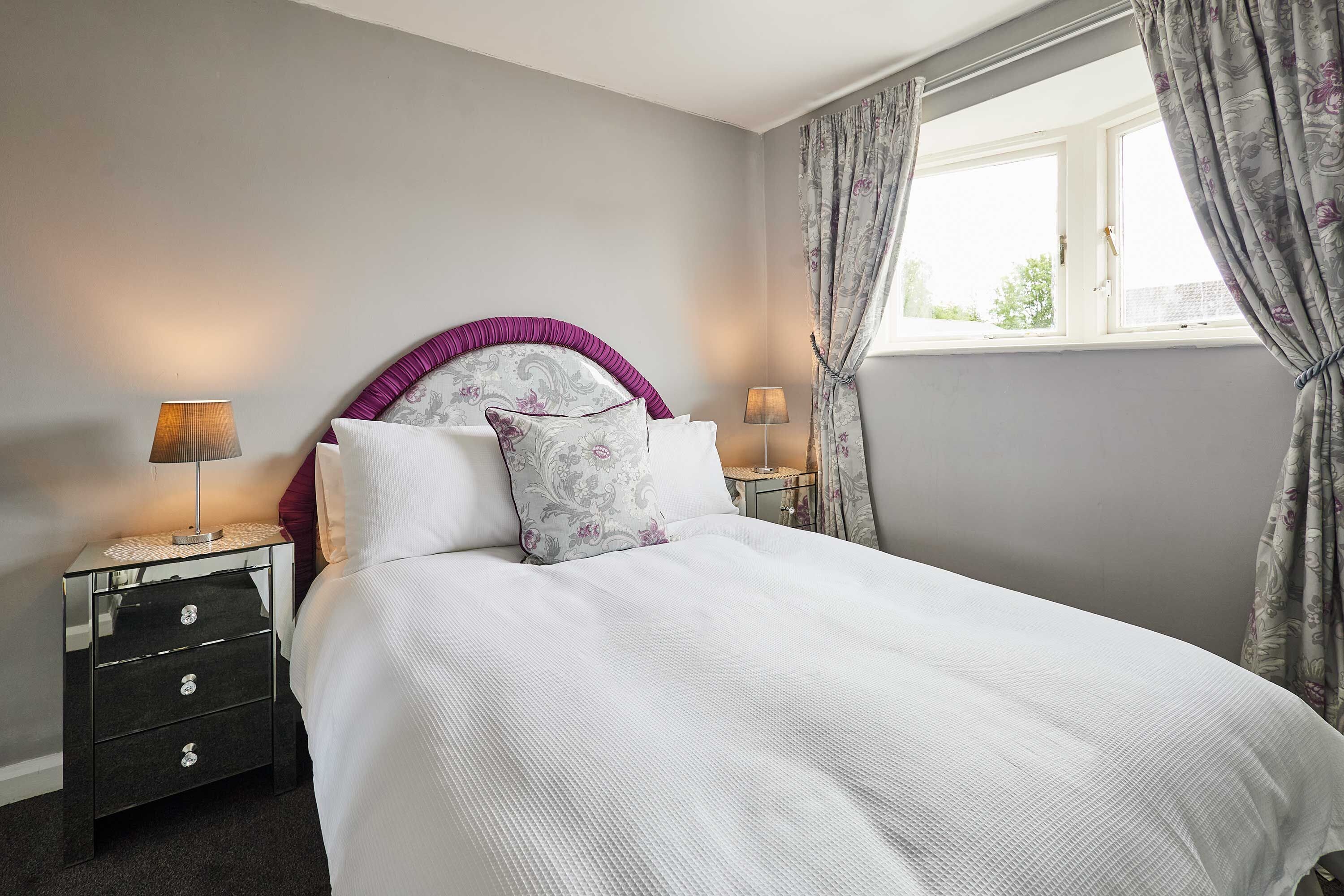A neatly made bed with a headboard, two bedside lamps, a nightstand, and a window with curtains.