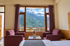 A hotel room with a bed, two armchairs, a small table with a vase, and a view of mountains and a village through large windows. - Echo Homestay (Manali)