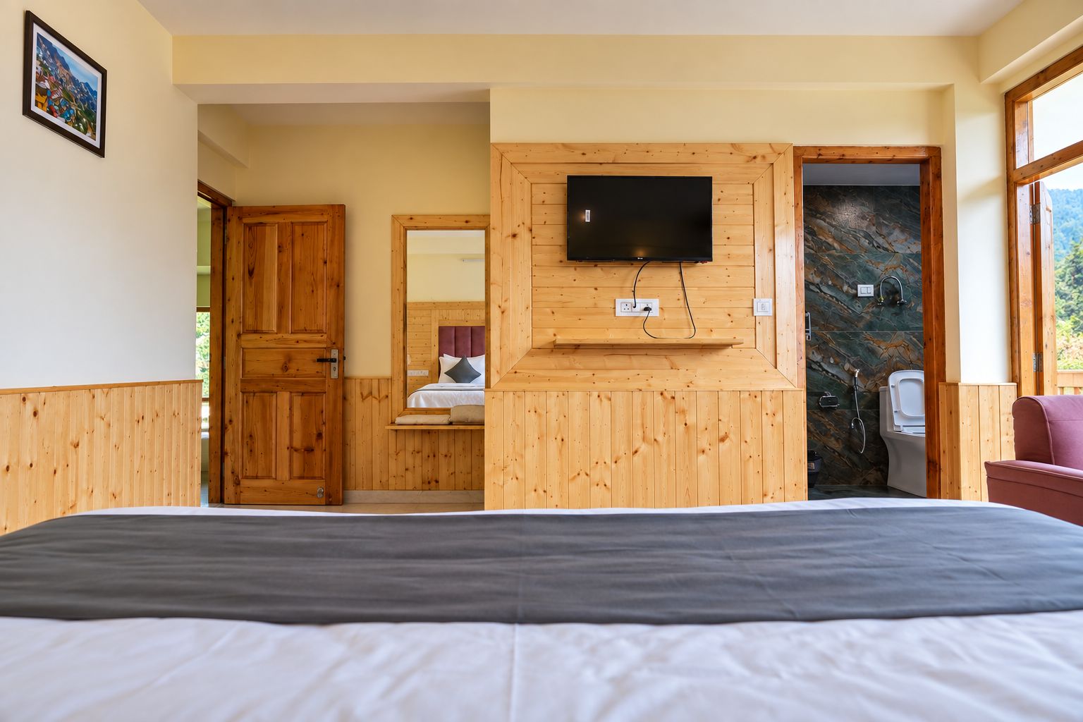 A hotel room with a bed, a television mounted on the wall, wooden paneling, a bathroom with a shower, and a view of the outdoors.