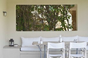 A patio with a white sofa and dining table, a wall-mounted light, and a view of dense foliage. - Villa Paradiso - Your Summer Sanctuary in Sani (Sane)