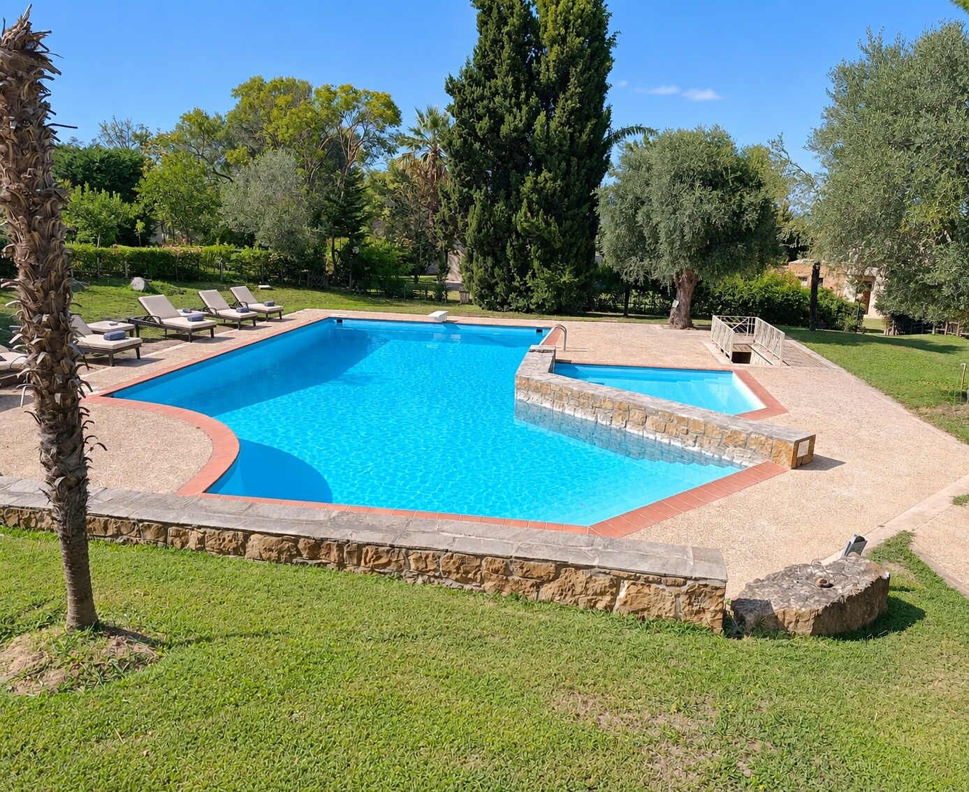 A rectangular swimming pool with a stone border, surrounded by grass and trees.