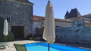 A swimming pool with lounge chairs and umbrellas, surrounded by stone buildings with tiled roofs.