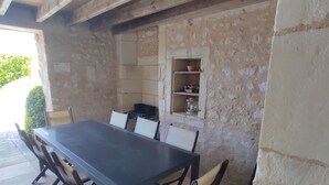 An outdoor dining area with a long table and chairs, a stone wall, and a shelf with kitchenware.