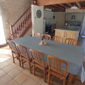 A dining area with a long table, wooden chairs, and a kitchen in the background.