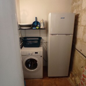A compact kitchen with a washing machine, a refrigerator, and a shelf with dishes and a blue container.