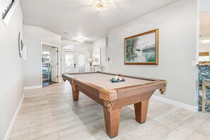 A pool table in a room with a wooden floor, a painting on the wall, and a door leading to another room. - Bees Bungalow - Lux Waterfront Pool & Spa w/ Dock & Lift - 2+ Acres - 360° Views (Matlacha)