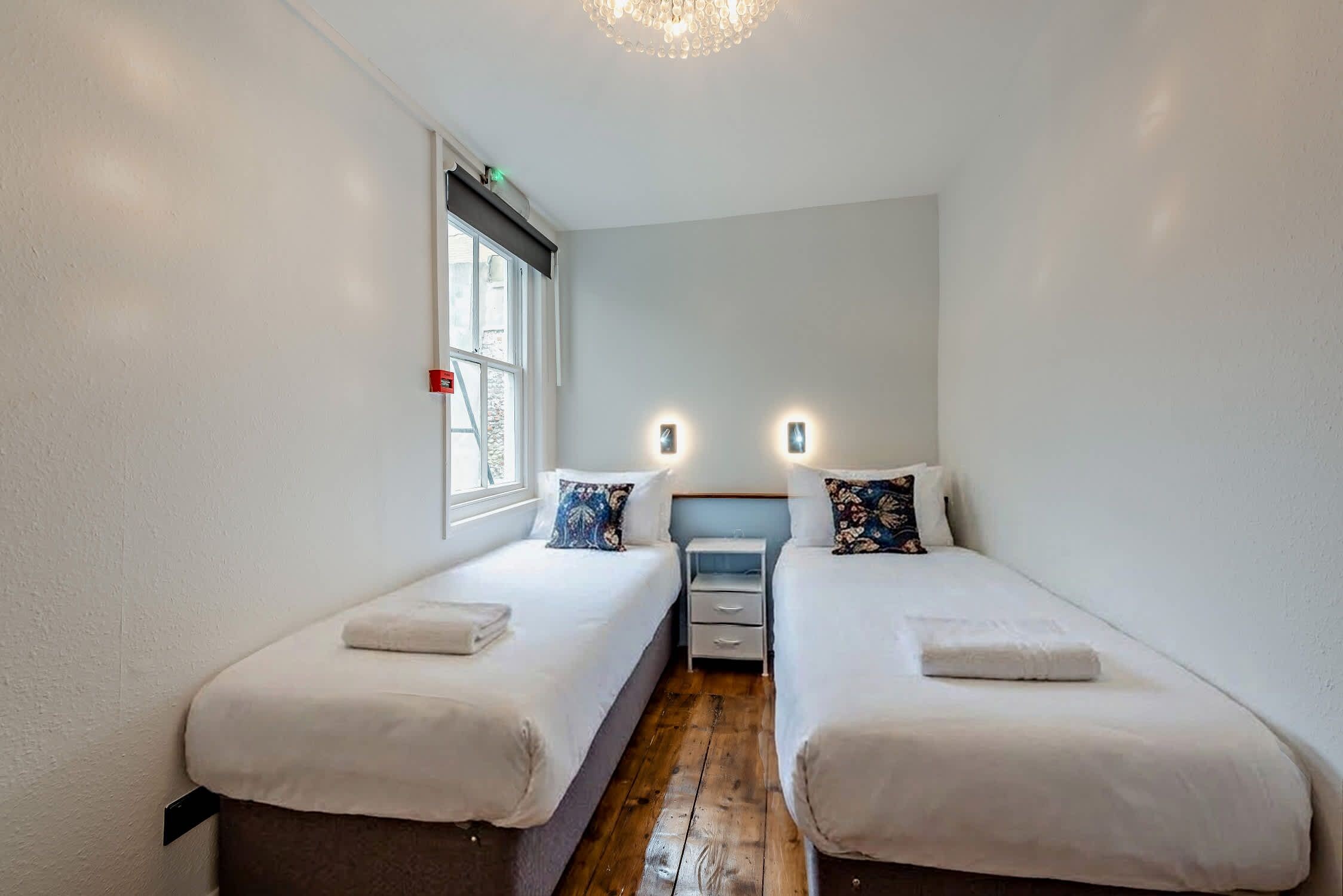 A small, narrow room with two single beds, a window, and a wooden floor.