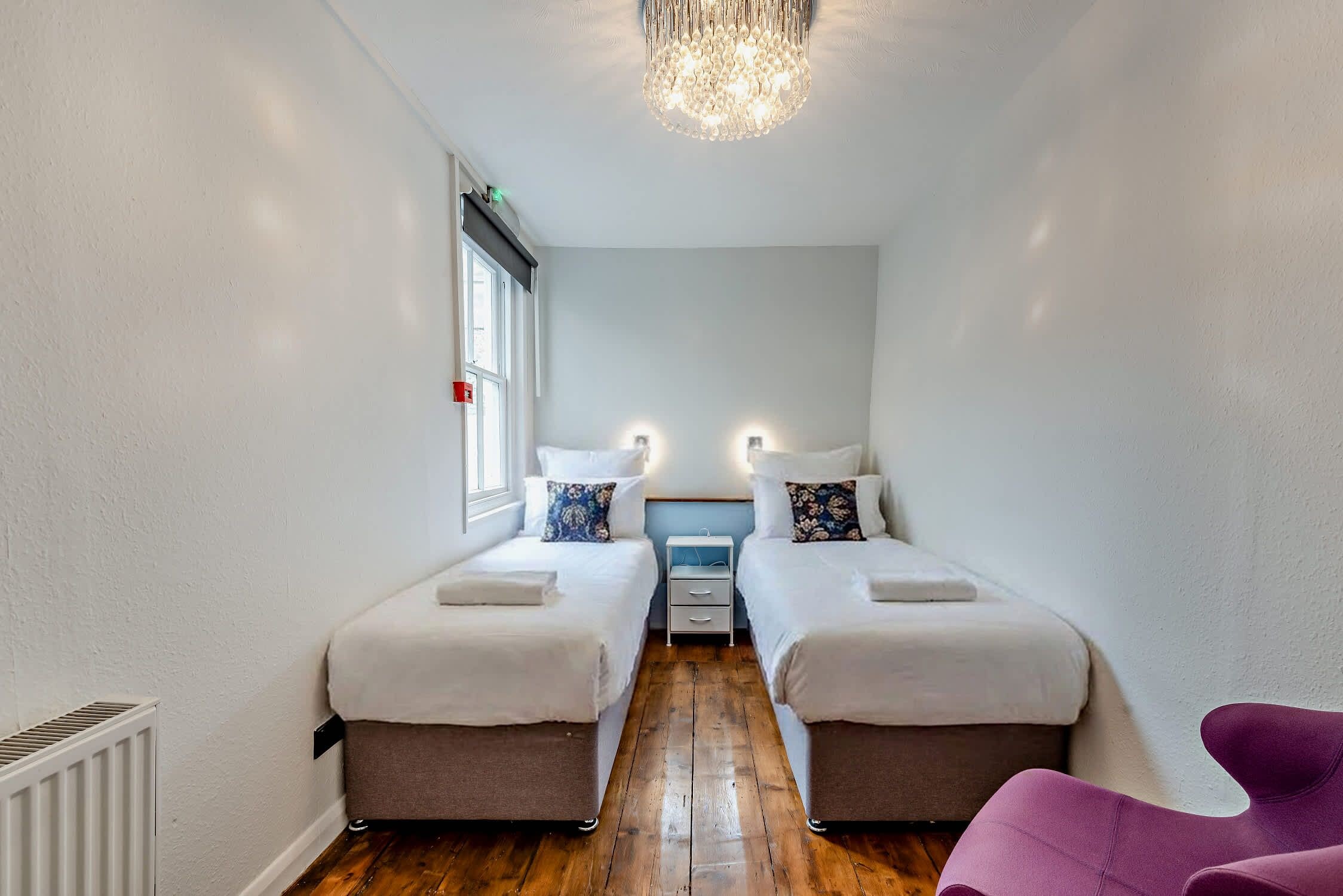 A narrow hotel room with two beds, a small bedside table, a chair, and a radiator.