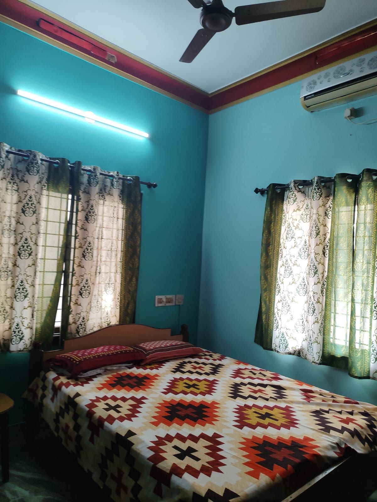 Ein Zimmer mit türkisfarbenen Wänden, einem Bett mit einem gemusterten Überwurf, Vorhängen mit Blumenmuster, einem Deckenventilator und einer Klimaanlage.
