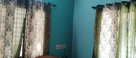 Una habitación con paredes color turquesa, una cama con colcha estampada, cortinas con diseño floral, un ventilador de techo y un equipo de aire acondicionado.