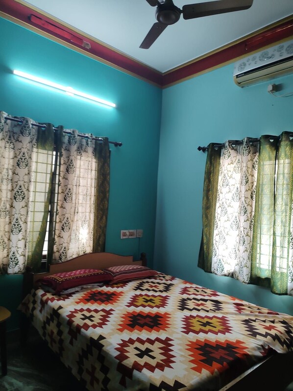 A room with teal walls, a bed with a patterned bedspread, curtains with a floral design, a ceiling fan, and an air conditioning unit.