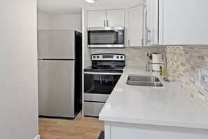 A compact kitchen with stainless steel appliances, including a refrigerator, microwave, and oven. - Ground Floor Beachfront Condo. Heated Pool. Shuffleboard. Perfect Location. No Car Necessary! (Treasure Island)