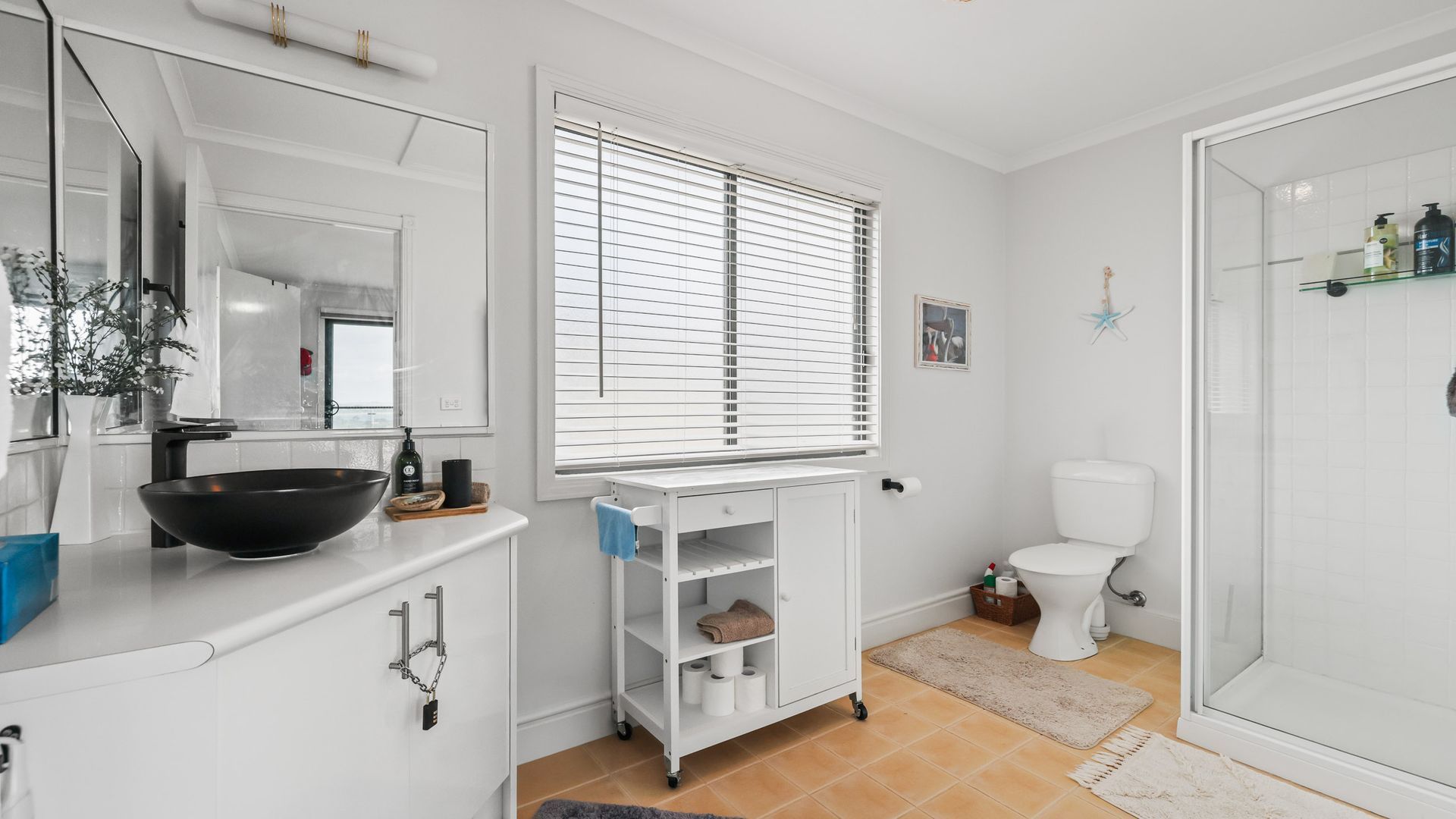 A bathroom with a white sink, mirror, and toilet. There is a window with blinds, a shower area, and a small shelf with toiletries.