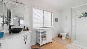 A bathroom with a white sink, mirror, and toilet. There is a window with blinds, a shower area, and a small shelf with toiletries. - Panorama Bay Escape San Remo (San Remo)